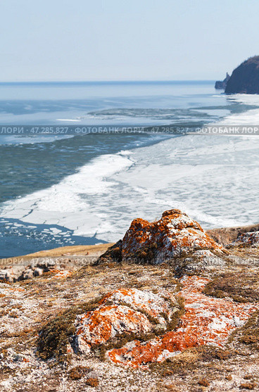 Baikal Lake in May. View from rocky shore of Olkhon Island to ice drift and to Cape Khoboy in distance. Beautiful spring landscape with stones covered with red lichens. Natural background. Changing seasons. Стоковое фото, фотограф Виктория Катьянова / Фотобанк Лори