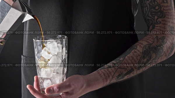 Barista with tattoo pours freshly made coffee into a glass with ice around a dark background. 4K UHD video, 240fps, 2160p.. A man with a tattoo pours coffee in a glass with ice around a dark background. 4K UHD video, 2160p. Стоковое фото, агентство Ingram Publishing / Фотобанк Лори