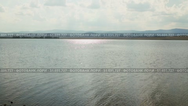 Bashkortostan, Russian Federation - August 07, 2025: Serene lake view reflecting sunlight with distant mountains under a cloudy sky, creating a tranquil natural landscape for relaxation and inspiration. Редакционное видео, видеограф Ирина Ручушкина / Фотобанк Лори
