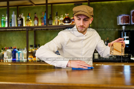 Bearded man in white shirt and cap removing napkin holder and wiping counter with cloth while working in bar in evening. Bartender cleaning counter in a nice pub. Стоковое фото, агентство Ingram Publishing / Фотобанк Лори