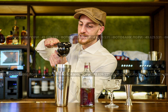 Bearded man in white shirt and cap squeezing lemon juice over shaker with cocktail while working behind counter in pub. Male barkeeper squeezing lemon into cocktail in a pub. Стоковое фото, агентство Ingram Publishing / Фотобанк Лори