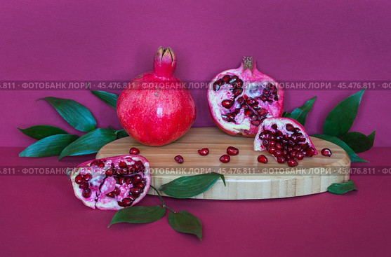 Beautiful still life with ripe pomegranates and pomegranate tree leaves on a wooden podium on a burgundy background. Advertisement of natural organic seasonal fruits with a high content of vitamins. Стоковое фото, фотограф Виктория Катьянова / Фотобанк Лори