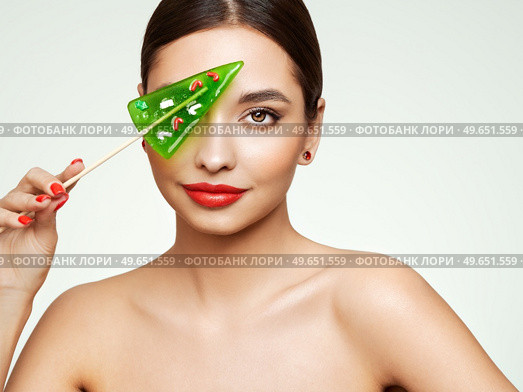 Beauty model girl eating Christmas tree shape lollipop.  Beautiful young woman with clean fresh skin. Girl beauty face care. Facial treatment. Cosmetology, beauty and spa. Red nails manicure. Стоковое фото, агентство Ingram Publishing / Фотобанк Лори