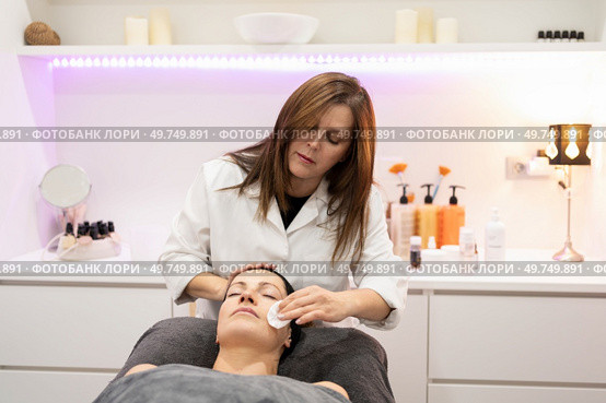 Beauty specialist removing cosmetic cream on face of female customer with skin care cotton pad while working in modern salon. Beautician cleaning face of woman with cotton pad in beauty salon. Стоковое фото, агентство Ingram Publishing / Фотобанк Лори