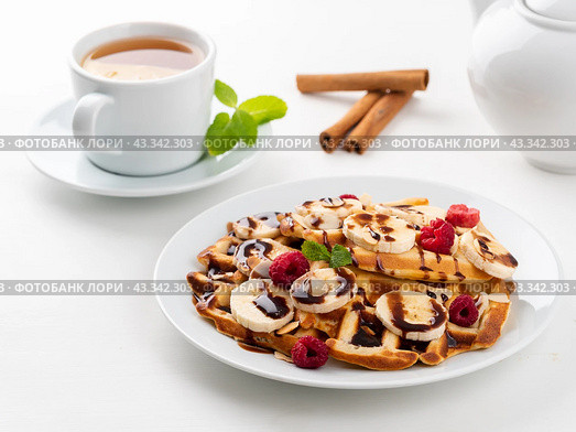 Belgian waffles with raspberries, banana, chocolate syrup. Breakfast with tea on white background. Стоковое фото, фотограф Natalia Gdovskaia / Фотобанк Лори