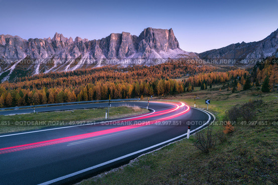 Blurred car headlights on winding asphalt road in mountains at sunset in autumn. Spectacular landscape with light trails, forest, rocks and purple sky at night in fall. Car driving on roadway. Стоковое фото, агентство Ingram Publishing / Фотобанк Лори