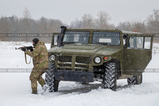 Боец в камуфляже с автомтом Калашникова занимает позицию у бронеавтомобиля "Тигр" снежным днем. Военно-исторический парк "Стальной десант", Красное село (2023 год). Редакционное фото, фотограф Виктор Карасев / Фотобанк Лори