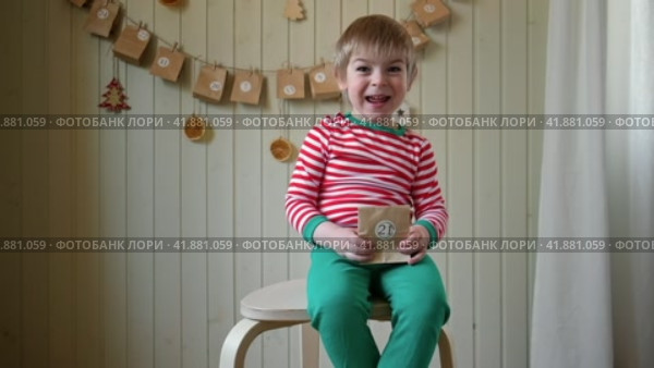 Boy in pajamas, Santa cap eating sweet candy on paper advent calendar with presents background. Little happy child with gift of handmade Christmas calendar on wall. Celebrating at home Xmas tradition. Стоковое видео, видеограф Марина Демидюк / Фотобанк Лори