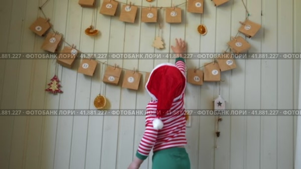Boy in pajamas, Santa cap eating sweet candy on paper advent calendar with presents background. Little happy child with gift of handmade Christmas calendar on wall. Celebrating at home Xmas tradition. Стоковое видео, видеограф Марина Демидюк / Фотобанк Лори