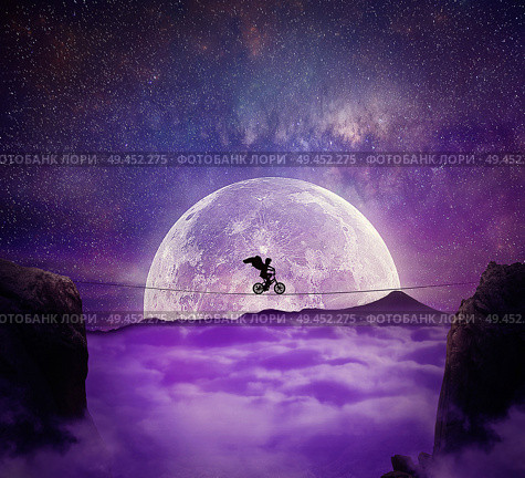 Boy with angel wings balance on a wire over a chasm riding a bicycle. Self overcoming and risk taking concept. Full moon night background over the clouds. Стоковое фото, агентство Ingram Publishing / Фотобанк Лори