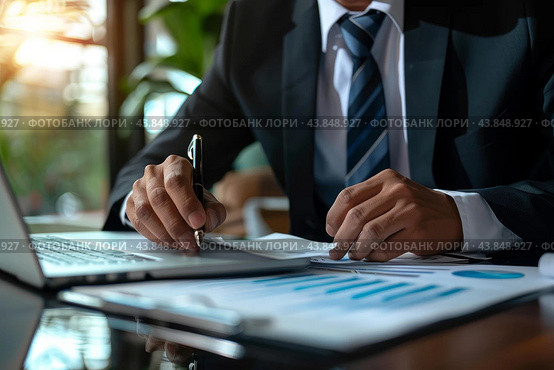 Businessman analyzing financial documents and using laptop in office. Стоковое AI-изображение, AI-художник Иван Рябоконь / Фотобанк Лори