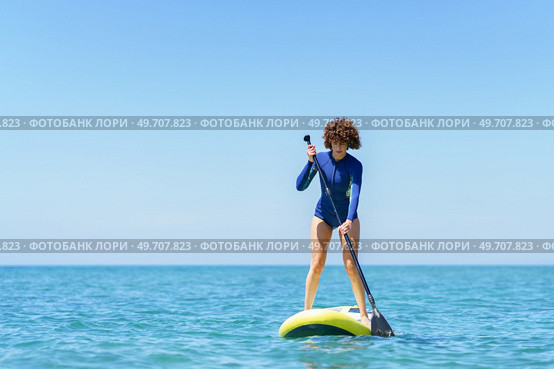 Careful female surfer in wetsuit standing on SUP board and paddling blue sea against cloudless blue sky in sunny summer day. Curly hair woman with paddleboard in sea. Стоковое фото, агентство Ingram Publishing / Фотобанк Лори
