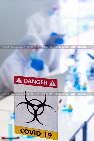 Caution danger Signage of COVID-19 coronavirus in front of Laboratory room with background of Scientists working and researching vaccine for ncov-19 virus for world pandemic situation. Стоковое фото, агентство Ingram Publishing / Фотобанк Лори