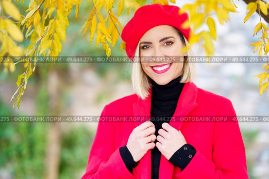 Charismatic young happy female, with blond hair and makeup in elegant red beret and coat smiling and looking at camera standing under tree with yellow leaves in autumn park. Joyful trendy lady smiling at camera in autumn garden. Стоковое фото, агентство Ingram Publishing / Фотобанк Лори