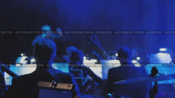 Часть оркестра во время выступления или репетиции на сцене с дирижером. Вид сзади показывает спины музыкантов и инструменты под эффектным сценическим освещением. Стоковое видео, видеограф Воловодов Владимир / Фотобанк Лори