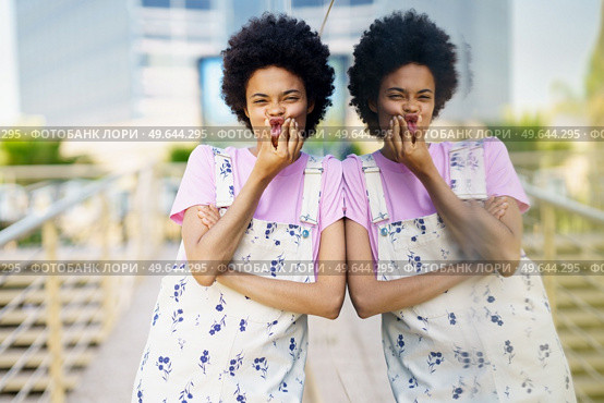 Cheerful African American female touching cheeks while making funny face and looking at camera near building with glass wall in city against blurred background. Positive black woman grimacing near building. Стоковое фото, агентство Ingram Publishing / Фотобанк Лори