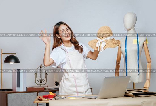 Cheerful Asian young female designer wearing eyeglasses doing fashion design on beautiful sombrero in studio, full of tailoring tools  with mannequin and equipment on the desk. Стоковое фото, агентство Ingram Publishing / Фотобанк Лори