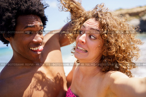 Cheerful black man with Afro hair and tongue out touching curly hair of girlfriend making funny grimace on sunny day near sea. Selfie of funny diverse couple making faces on beach. Стоковое фото, агентство Ingram Publishing / Фотобанк Лори