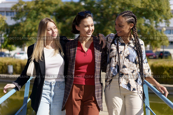 Cheerful multiracial female friends looking at each other while standing together on bridge over river in city on summer day. Positive diverse women on footbridge in city. Стоковое фото, агентство Ingram Publishing / Фотобанк Лори