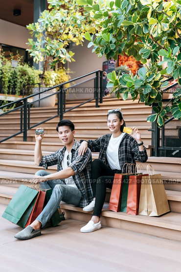 Cheerful Young couple holding multiple paper shopping bag on stairs of large shopping mall and show credit card in hand,  Lifestyle and shopping concept, copy space. Стоковое фото, агентство Ingram Publishing / Фотобанк Лори