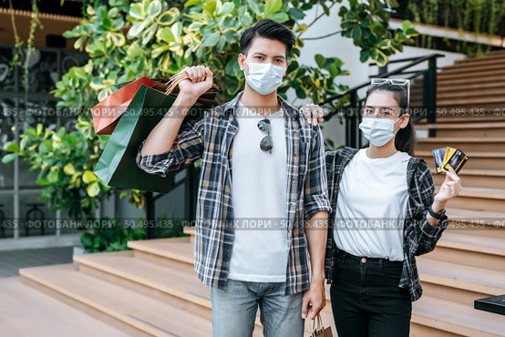 Cheerful Young couple in mask holding multiple paper shopping bag going down stairs of large shopping mall and pretty woman show credit card in hand,  Lifestyle and shopping concept, copy space. Стоковое фото, агентство Ingram Publishing / Фотобанк Лори
