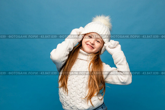 Child girl in a white sweatshirt and knitted hat with pompom on a blue background. The concept of children's emotions, joy and delight. Advertising clothing and sales. Ski resort vacation, copy space. Стоковое фото, фотограф Ирина Михайличенко / Фотобанк Лори