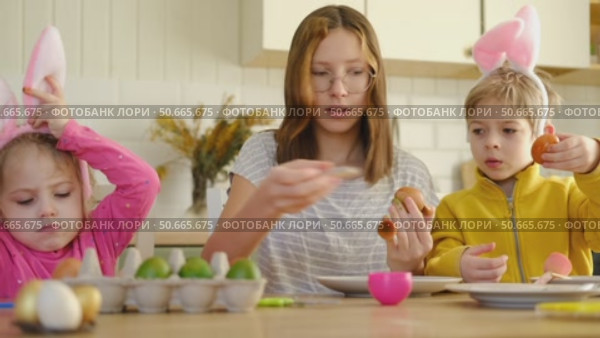 Children siblings painting Easter egg sitting together at table. Easter Family traditions. Teen girl teaching happy little brother and sister to dye, decorate eggs with paints. Preparation to holidays. Стоковое видео, видеограф Марина Демидюк / Фотобанк Лори