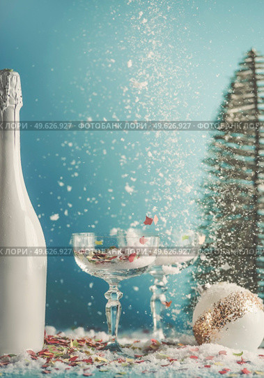 Christmas and New Year’s Eve background with champagne glasses and white champagne bottle. Christmas bauble and Christmas tree at blue background with snow and confetti. Front view. Стоковое фото, агентство Ingram Publishing / Фотобанк Лори