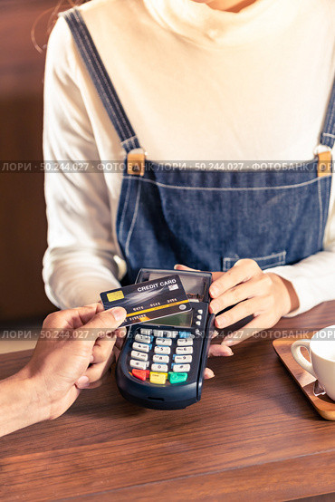 Close-up of asian customer using his credit card with contactless nfs technology to pay a barista for his coffee purchase at a cafe bar. Стоковое фото, агентство Ingram Publishing / Фотобанк Лори
