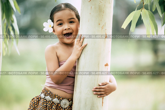 Close up, Portrait of little girl  in Thai traditional dress and put white flower on her ear, Stand and embrace trunk of the tree, laugh and looking at camera, copy space. Стоковое фото, агентство Ingram Publishing / Фотобанк Лори