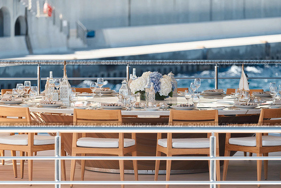 Close-up view of luxurious setting of the banquet table on an expensive mega yacht at sunny day, with awnings stretched over the deck to protect from the sun, wealth life, flowers and chairs. Стоковое фото, фотограф Vladimir Drozdin / Фотобанк Лори
