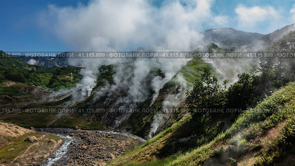 Columns of steam from hot springs rise above the slopes of the mountain. The stream flows in a hollow along a rocky bed. Blue sky. Kamchatka. Valley of Geysers. Стоковое фото, фотограф Вера Ивановна Кубарева / Фотобанк Лори