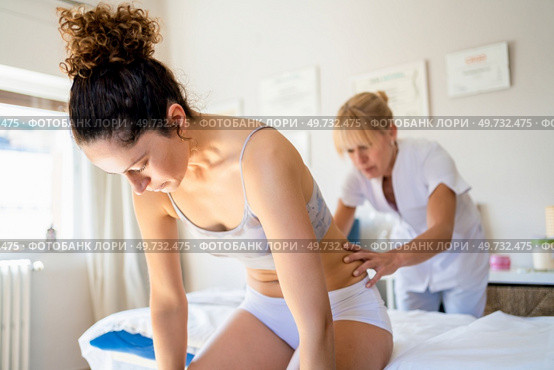 Concentrated masseuse looking down, while standing near spa bed and massaging lower back of young slim female sitting and bending forward during physiotherapy in osteopathy clinic. Focused masseuse massaging back of client in clinic. Стоковое фото, агентство Ingram Publishing / Фотобанк Лори