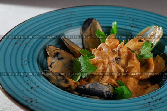 Cooked mussels in shells served with a rich spicy creamy sauce and fresh herbs in a rustic blue bowl on a light tablecloth, top-down view. Стоковое фото, фотограф Aleksandr Gavrilychev / Фотобанк Лори