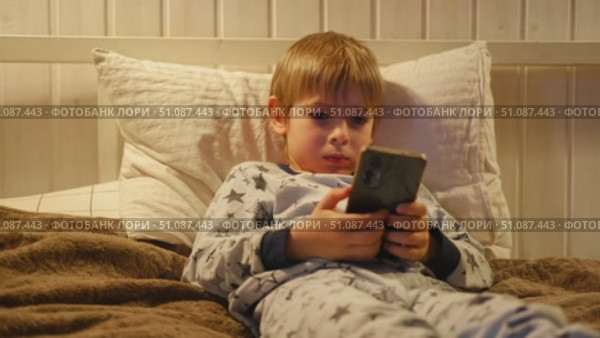 Cute kid boy in pajamas lying in bed watching smartphone, smiling. Children's screen time. Child plays games using phone at the evening or night. Social media addiction. Childhood, modern technology. Стоковое видео, видеограф Марина Демидюк / Фотобанк Лори