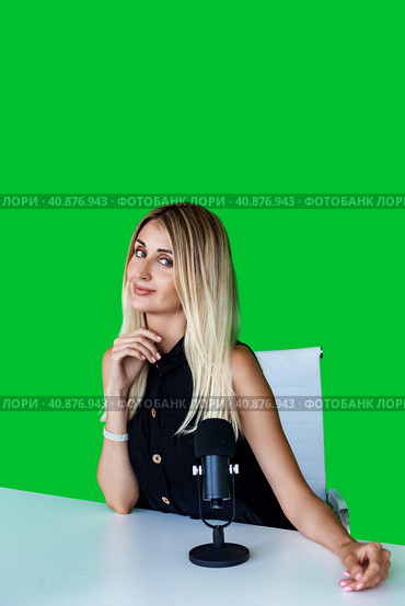 Cute young woman broadcaster sitting at desk with microphone at green backdrop, looking at camera. Portrait of female broadcaster posing in television studio. Tv production concept. Copy ad text space. Стоковое фото, фотограф Aleksandr Golubev / Фотобанк Лори