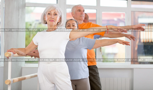 Dancing positive elderly women and men engaged in group class perform exercise near ballet barre, standing in 2nd position of ballet stance in dance studio. Стоковое фото, фотограф Яков Филимонов / Фотобанк Лори
