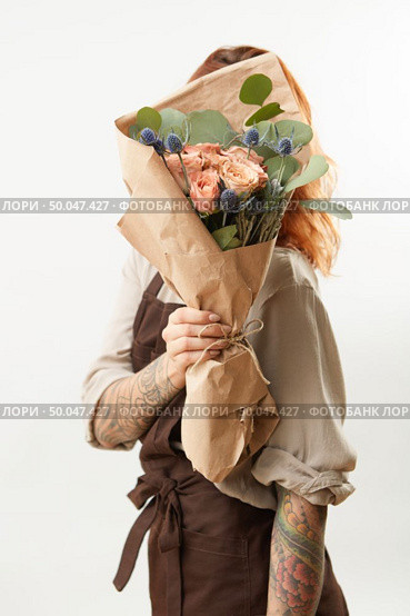 Delicate flowers coral living roses and eryngium as a beautiful bouquet hide girl’s face and ginger hair on a gray background, place for text. Concept of Mother’s or Woman’s Day.. Girl with red hair hide her face big fresh bunch from natural roses and green leaves on a light background. Copy space. Congratulation card. Стоковое фото, агентство Ingram Publishing / Фотобанк Лори