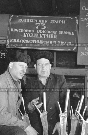 Двое мужчин сидят рядом с производственным помещением и надписью "Коллективу драги 73 присвоено высокое звание коллектива коммунистического труда", Якутия, март 1961 год. Редакционное фото, фотограф Варфоломей Тетерин / Фотобанк Лори