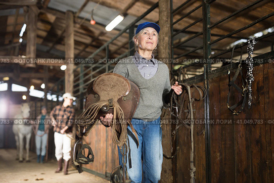 Elderly female rancher walking in stable with saddle in hands. Стоковое фото, фотограф Яков Филимонов / Фотобанк Лори