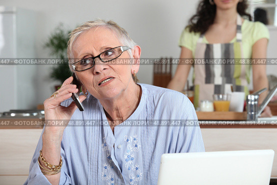 Elderly woman chatting on the phone whilst young helper works in the kitchen. Стоковое фото, агентство Ingram Publishing / Фотобанк Лори