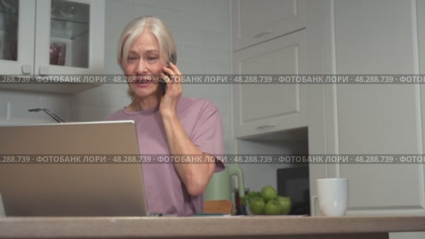 Elderly woman multitasking with phone and laptop in modern kitchen setting. Стоковое видео, видеограф Владимир Федоров / Фотобанк Лори