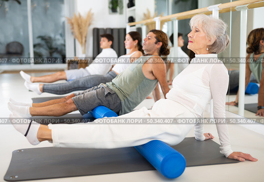 Elderly woman practicing pilates with roller on gray mat in gym room. Стоковое фото, фотограф Яков Филимонов / Фотобанк Лори