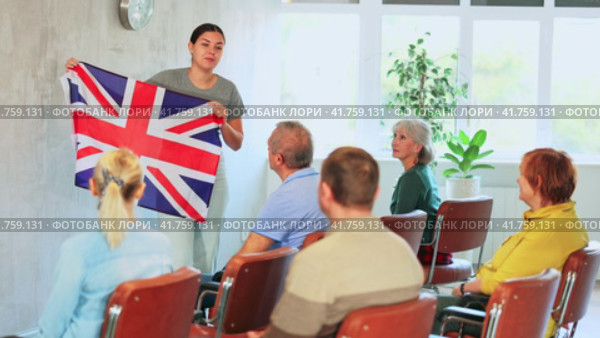 Emotional young female tutor showing national flag of United Kingdom while leading english language club for group of elderly people sitting in auditorium. Стоковое видео, видеограф Яков Филимонов / Фотобанк Лори