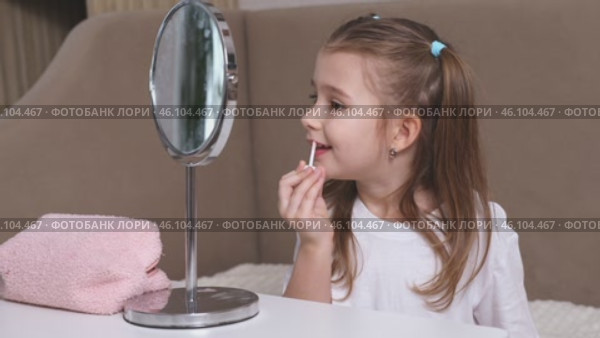 Fashion little girl in a white T-shirt is sitting at the dressing table in front of the mirror and applying makeup with lipstick. Advertising of children's cosmetics concept. Footage for kids blog. Стоковое видео, видеограф Ирина Михайличенко / Фотобанк Лори