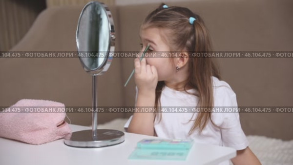 Fashion little girl in a white T-shirt is sitting at the dressing table in front of the mirror and applying makeup with eye shadow. Advertising of children's cosmetics concept. Footage for kids blog. Стоковое видео, видеограф Ирина Михайличенко / Фотобанк Лори