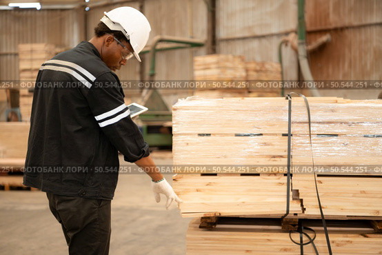 Female carpenter working in a woodworking factory, she is wearing a safety helmet and glasses.Checking the wooden planks coming out of the wood grinder. Стоковое фото, агентство Ingram Publishing / Фотобанк Лори