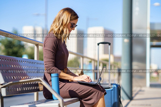 Female manager in stylish dress and sunglasses sitting on bench near suitcase and thermos and browsing netbook on station during business trip in city. Businesswoman using laptop on bench. Стоковое фото, агентство Ingram Publishing / Фотобанк Лори