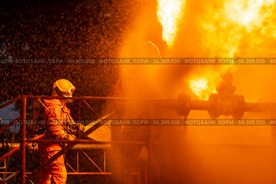 Firefighter using water fog type fire extinguisher to fighting with the fire flame from oil pipeline leak and explosion on oil rig and natural gas station. Firefighter and industrial safety concept. Стоковое фото, агентство Ingram Publishing / Фотобанк Лори