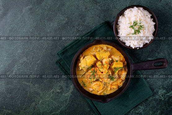 Fish curry with codfish, pollack and shrimps with coconut milk served in cast iron pan and bowl of rice on green table. Top view. Copy space. Стоковое фото, фотограф Оксана Чаюн / Фотобанк Лори